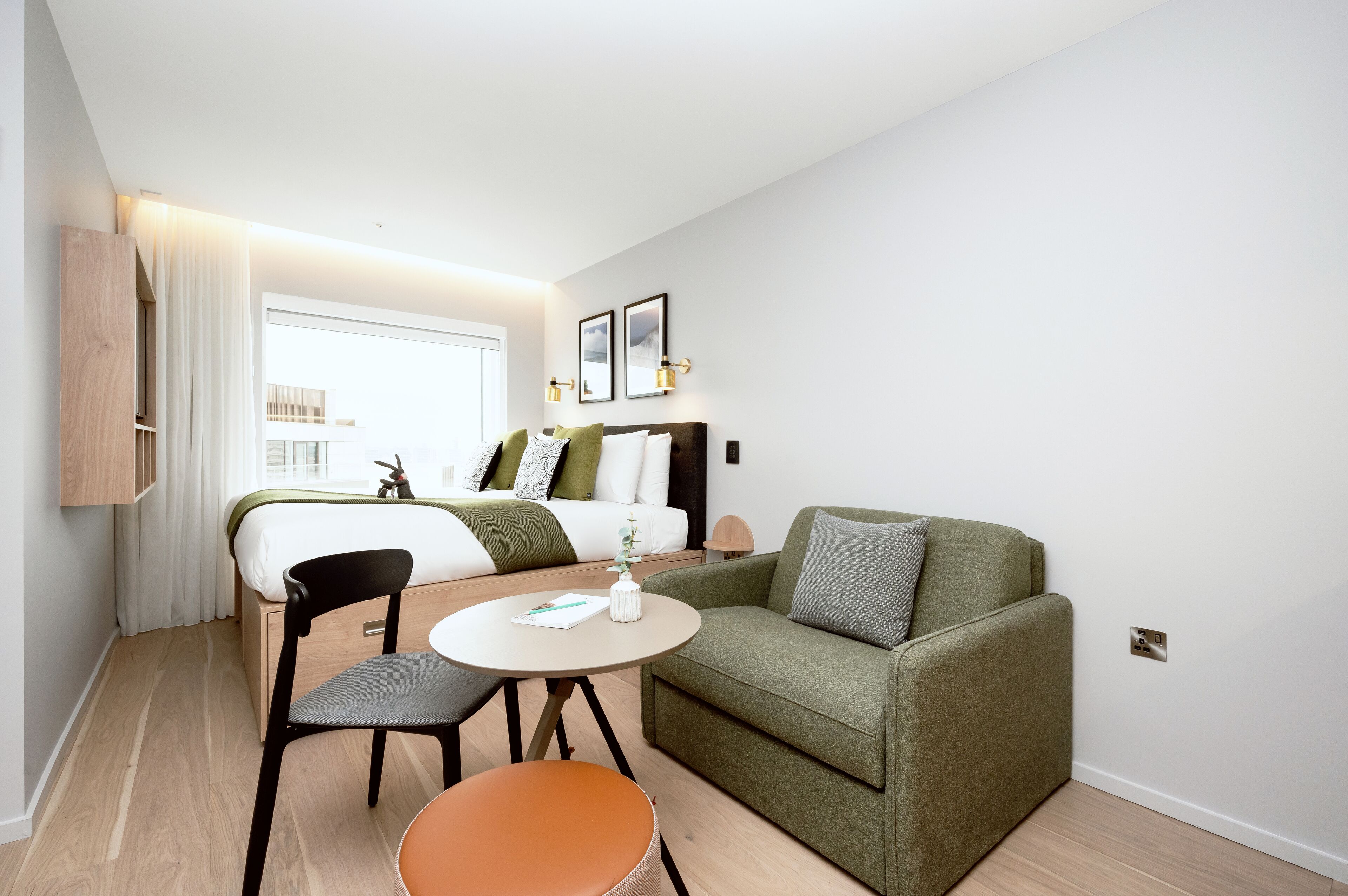 Een moderne hotelkamer met een bed, een groene bank, een ronde tafel met een stoel en een kleine tafel met een plant.