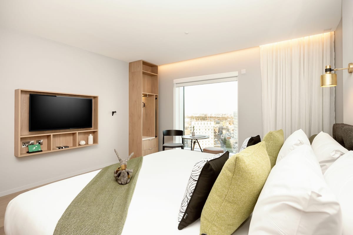 Une chambre d’hôtel moderne dotée d’un grand lit, d’un téléviseur à écran plat fixé au mur, d’une petite table avec des chaises et d’une grande fenêtre donnant sur la ville.
