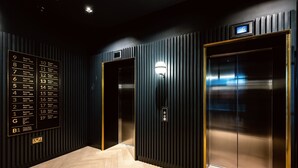 A hotel lobby with a directory on the left, two elevators, and a herringbone-patterned floor. - Wilde Aparthotels, London, Paddington (London)