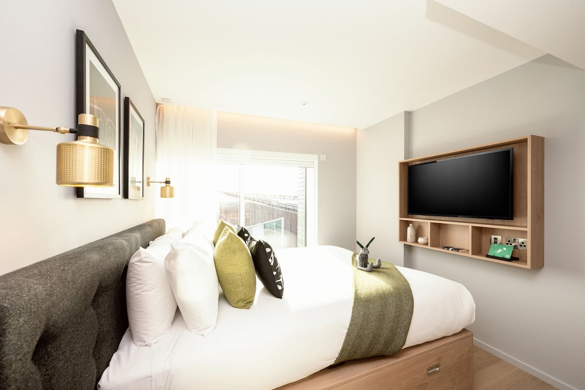 Une chambre d’hôtel moderne avec un grand lit, un téléviseur fixé au mur et une fenêtre donnant sur un immeuble.