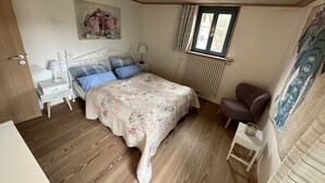 Ett sovrum med en säng, två sängbord, en stol, ett element och ett fönster med gardiner.