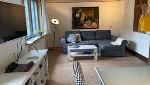 Ett vardagsrum med en grå soffa, ett soffbord, en golvlampa, en tavla på väggen och ett fönster med gardiner.