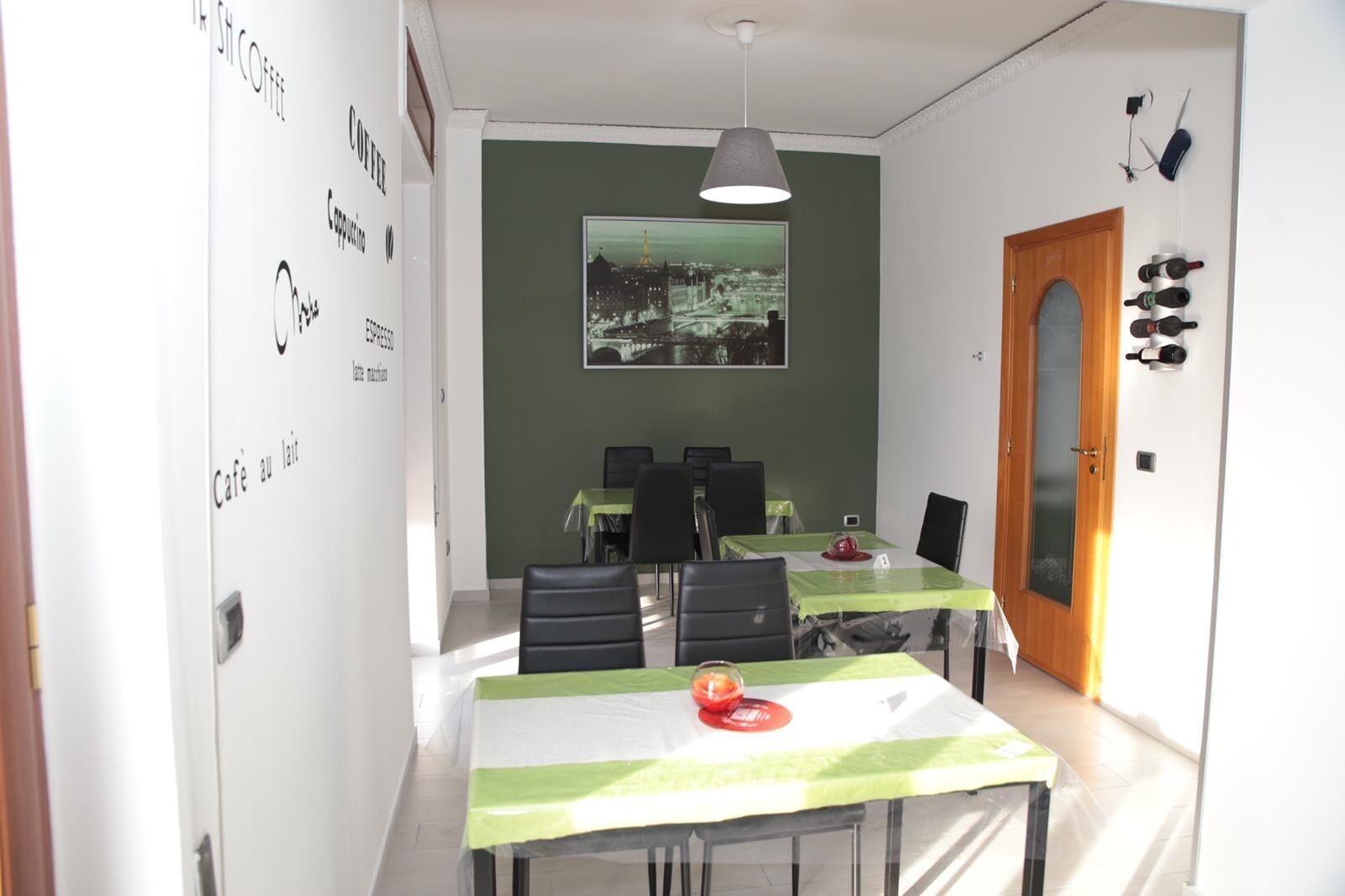 A dining area with tables and chairs, a painting on the wall, and a door with wine bottles.