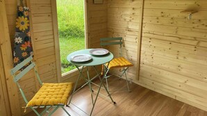 A small wooden room with a window, a table with plates, two chairs, a clock, and a wall-mounted shelf.