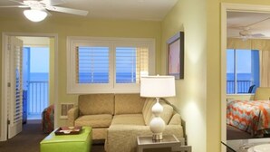 Ein Hotelzimmer mit Sofa, Hocker, einem Beistelltisch mit Lampe und Meerblick durch die Fenster.