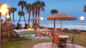 Eine Terrassenanlage im Freien mit Feuerschale, Liegestühlen und Sonnenschirmen, mit Blick auf einen Strand mit Palmen und dem Meer.