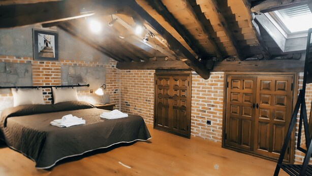 A rustic room with exposed wooden beams, a bed with a dark cover, a wooden dresser, and a skylight. - Casa Rural de Acogedor Estilo Castellano (Velayos)
