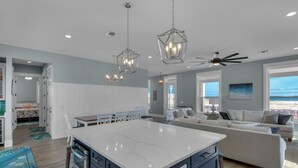 Ett rymligt kök med en stor köksö, vita skåp och blå detaljer. Här finns en matplats med ett vitt bord och stolar, och ett vardagsrum med en vit soffa och utsikt över havet.