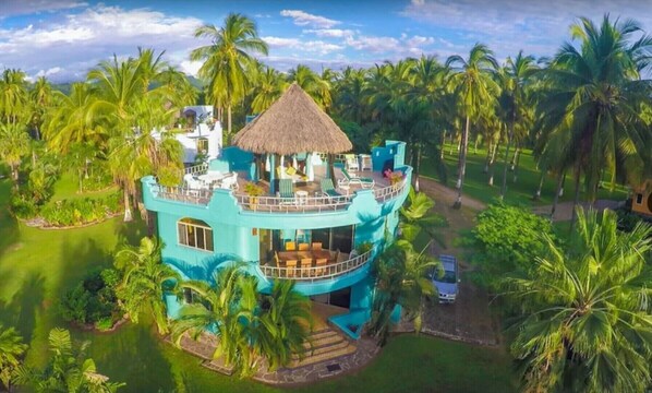 Experience the tropical ambiance of this multi-level resort, featuring a central building with a thatched roof and scenic views of palm trees and lush greenery.