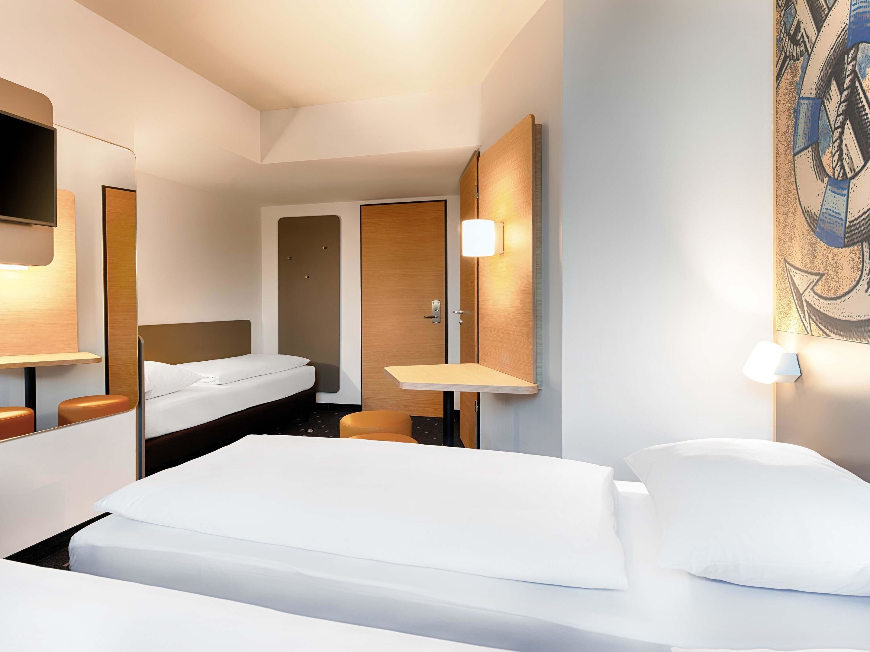 A hotel room with a bed, a desk, a chair, a mirror, and a wall-mounted TV.