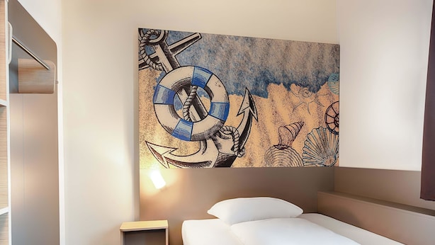 A hotel room with a bed, a small bedside table, a wall-mounted artwork, and a carpeted floor. - B&B Hotel Emden (Emden)