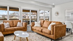 A spacious living room with two brown leather sofas, a glass coffee table, and a ceiling fan. Large windows offer a view of neighboring houses. - Modern Lakeview with All Ensuites, Entertainment Room, & Outdoor Fire Features (Branson)