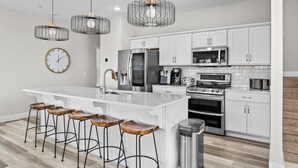 A modern kitchen with white cabinets, a central island with stools, stainless steel appliances, and geometric pendant lights. - Modern Lakeview with All Ensuites, Entertainment Room, & Outdoor Fire Features (Branson)