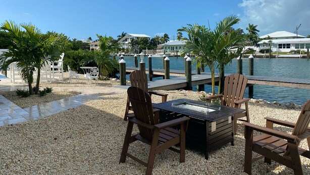 Une terrasse au bord de l’eau, avec des chaises et une table en bois, d’où l’on a vue sur une marina, avec des bateaux et des maisons en arrière-plan.