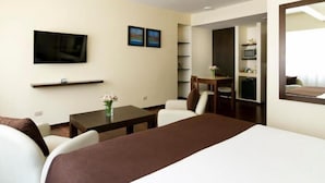 A hotel room with a bed, a TV, a desk with a chair, a small table with a vase, and a microwave in the kitchenette.