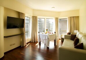 A hotel room with a flat-screen TV mounted on the wall, a dining table with a white tablecloth, a sofa, and large windows with curtains.