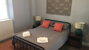 A neatly made bed with grey bedding and two red pillows, two bedside tables with lamps, a patterned wall hanging, and a window with blinds.