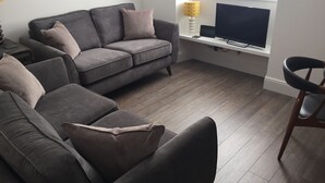A living room with a grey sofa, a wooden floor, a TV on a stand, and a window with blinds.