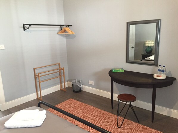A hotel room with a bed, a small table with a chair, a mirror, a hanging rack, and a trash bin.