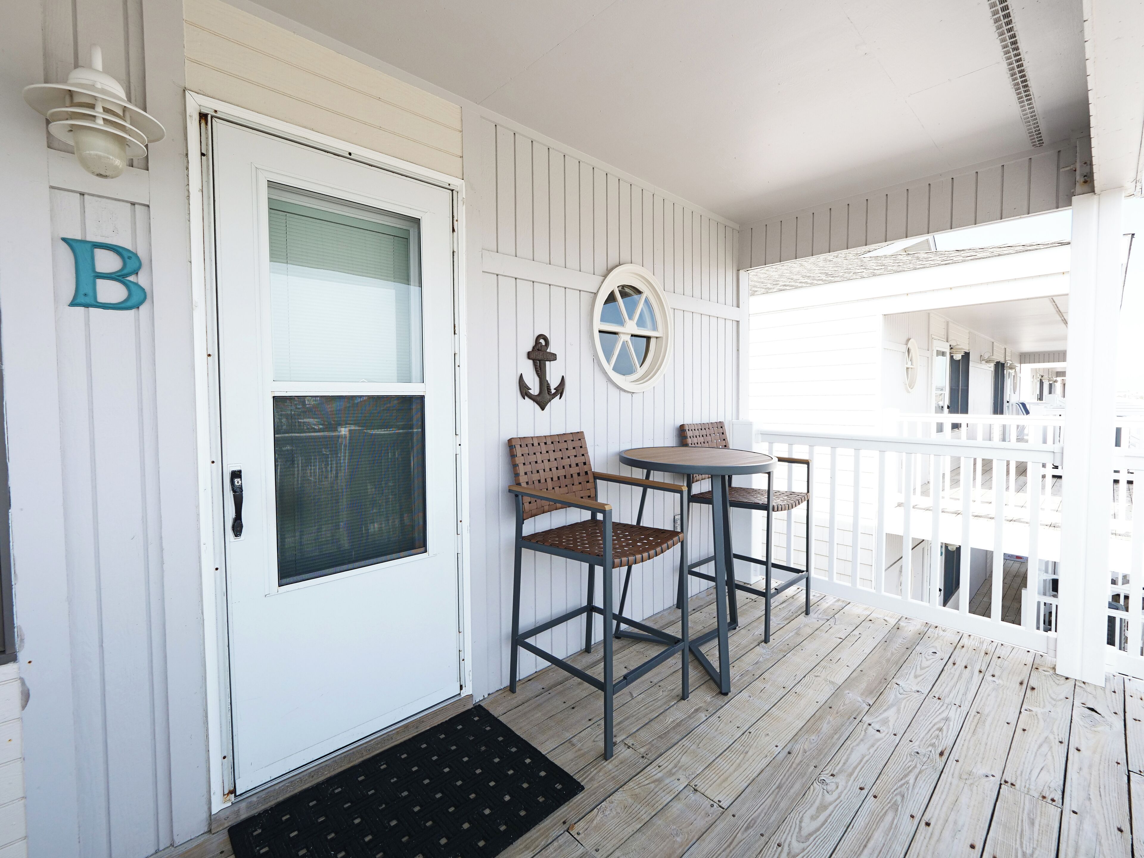 A wooden porch with a white door, two wicker chairs, a small round table, and a black doormat.