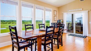 A dining area with a wooden table and chairs, large windows with blinds, and a view of a green landscape. - "Cool Fish" - 2nd Row Home w/ Private Pool, 5 Bedrooms / 4.5 Bathrooms (Holden Beach)
