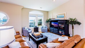 A living room with a large window, leather sofas, a coffee table, a TV, and a bookshelf. - "Cool Fish" - 2nd Row Home w/ Private Pool, 5 Bedrooms / 4.5 Bathrooms (Holden Beach)
