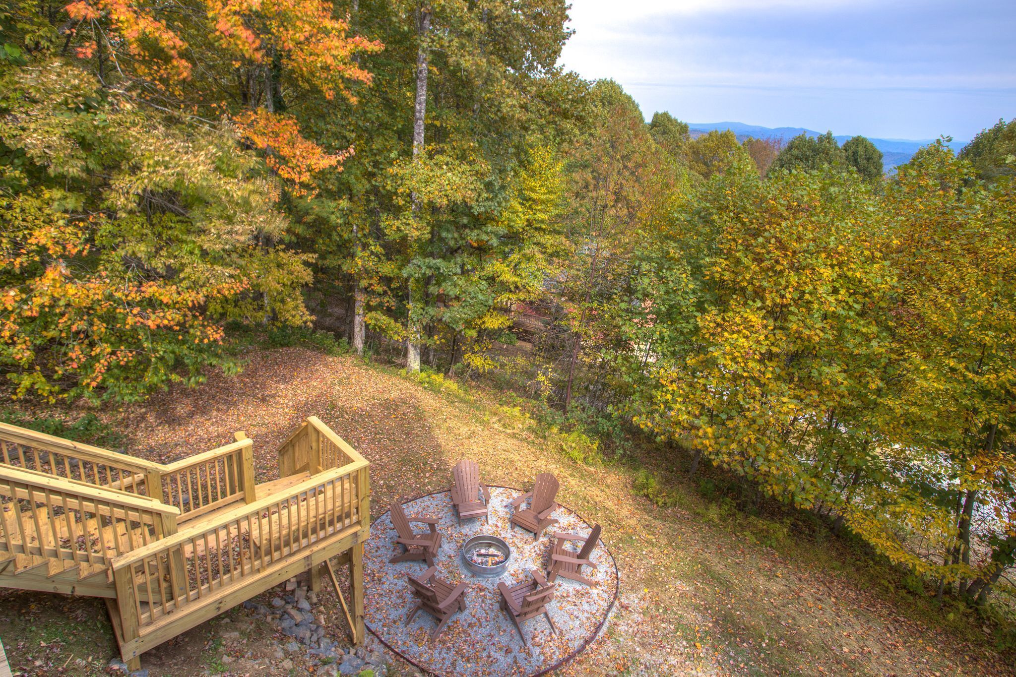 Un espace extérieur circulaire aménagé avec des chaises Adirondack et un foyer, entouré d’une terrasse en bois et d’une colline boisée.