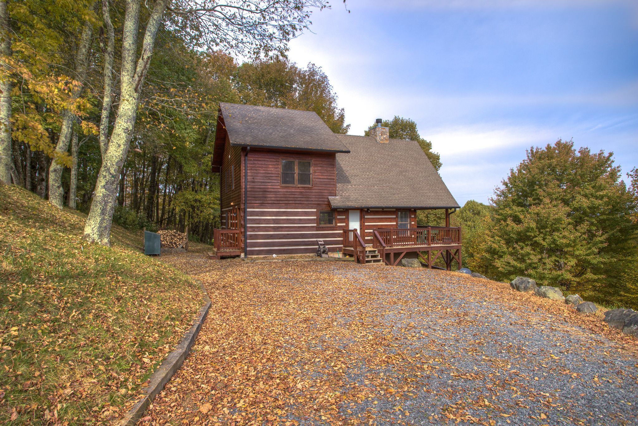 A rustic cabin with a wooden exterior, a porch, and a gravel driveway surrounded by trees.