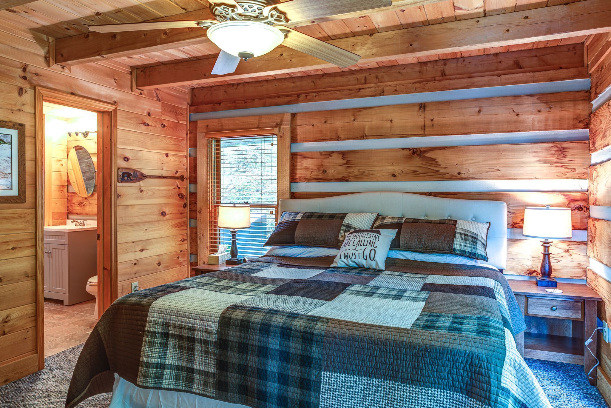 A cozy log cabin bedroom with a large bed, a ceiling fan, and a window with blinds.