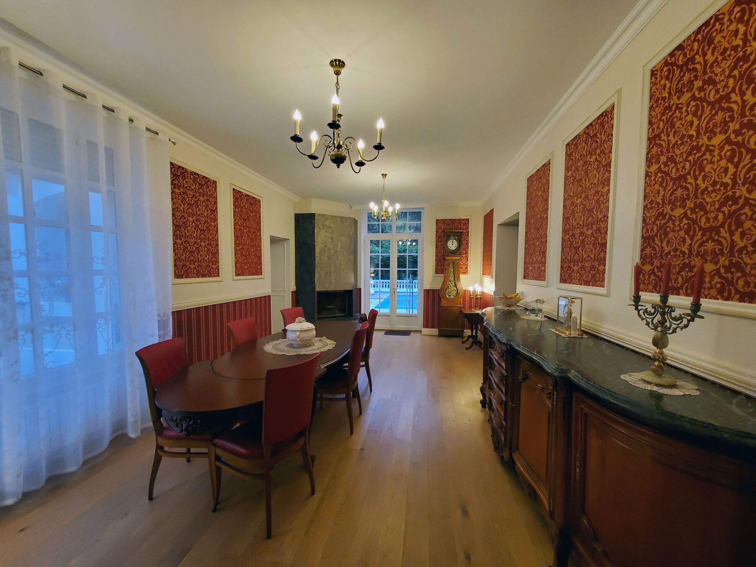 A dining room with a wooden table and red chairs, a chandelier, and a clock on the wall.