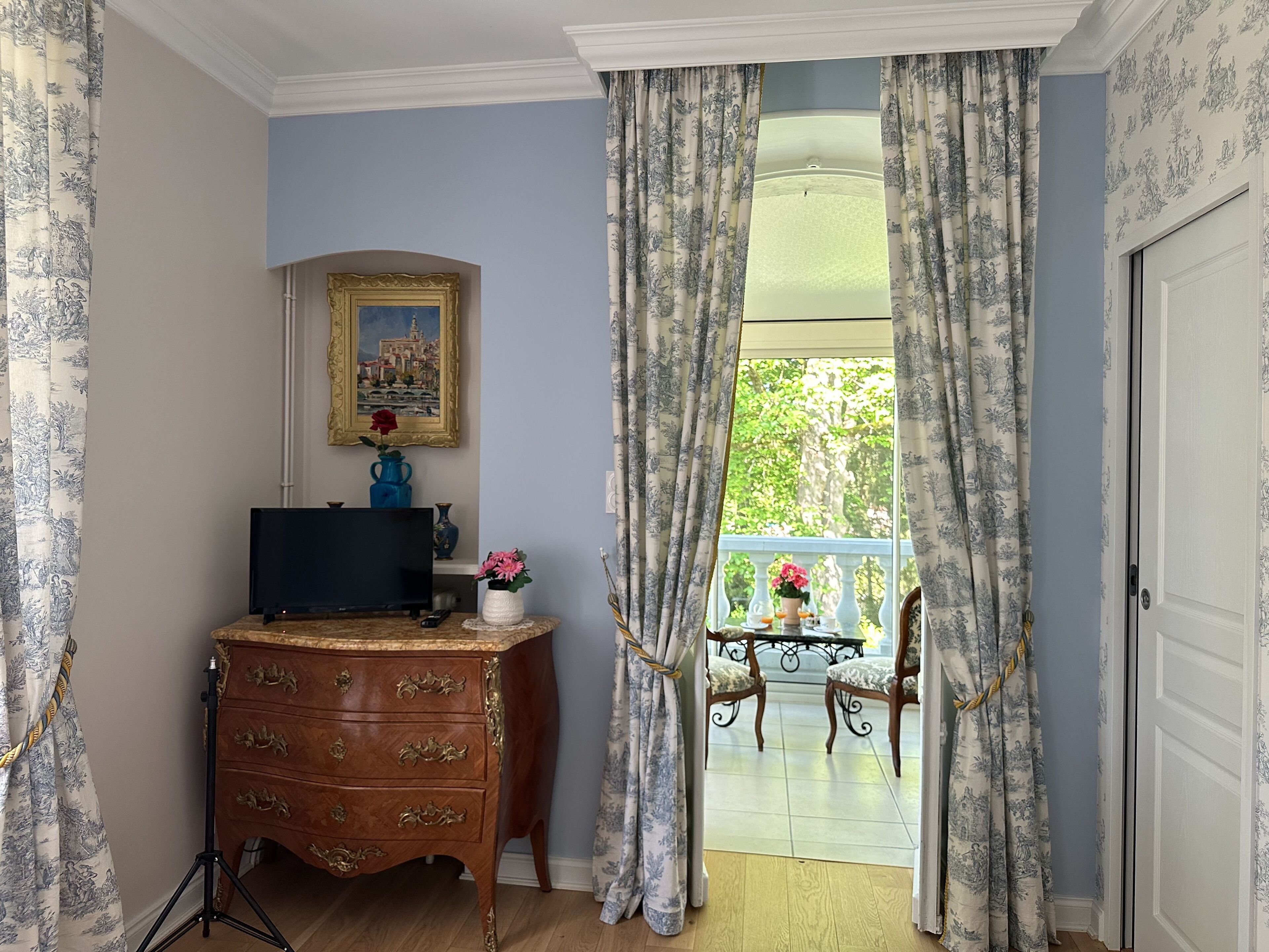 A room with a wooden dresser, a framed painting, and a view of a balcony with chairs and a table.