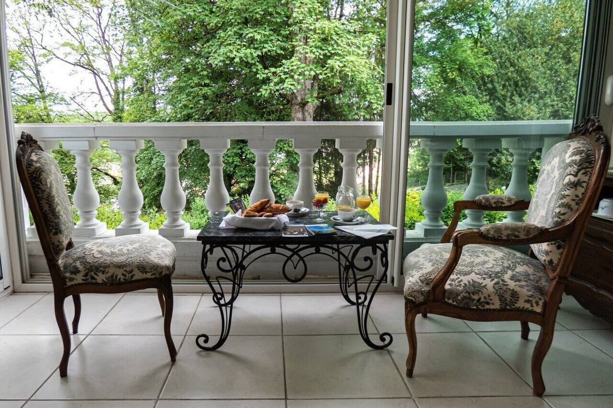 A conservatory with two upholstered chairs and a table, surrounded by a white balustrade, overlooking a lush green garden.
