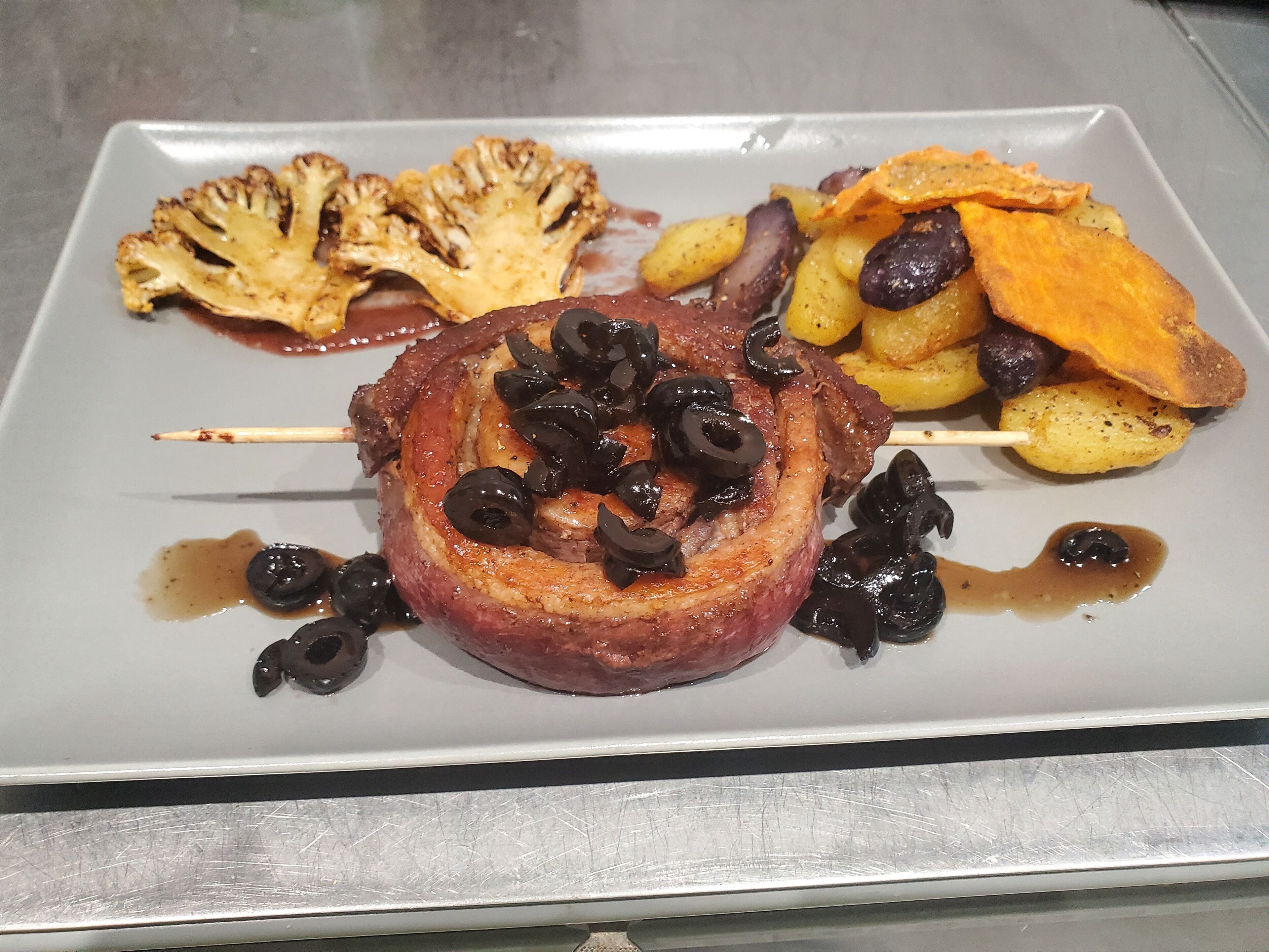 Un plato con un pincho de carne, patatas asadas y una guarnición que parece una preparación de verduras.