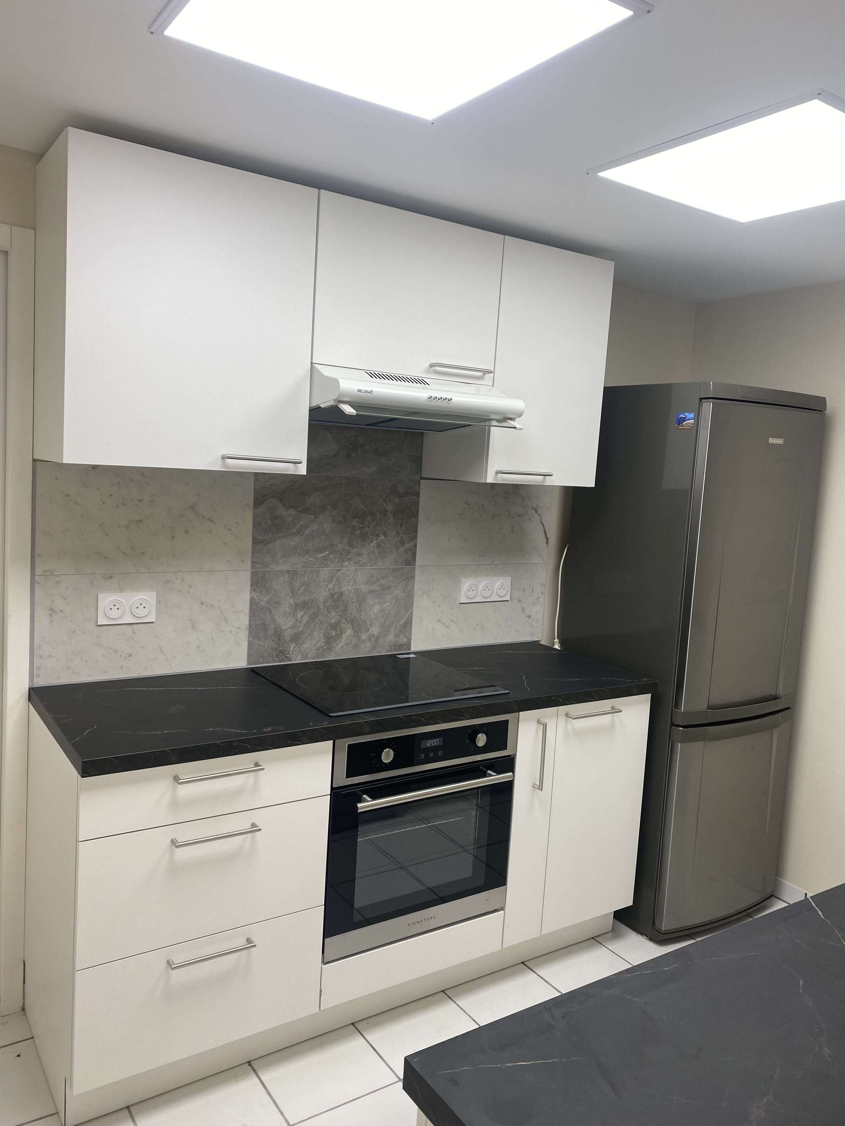 A modern kitchen with white cabinets, a black countertop, a built-in oven, a stainless steel refrigerator, and a skylight above.