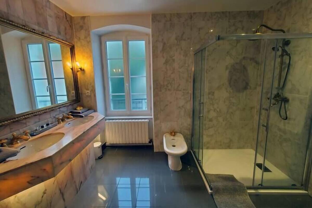 A bathroom with a marble vanity, a glass shower enclosure, a bidet, and a window with frosted glass panels.