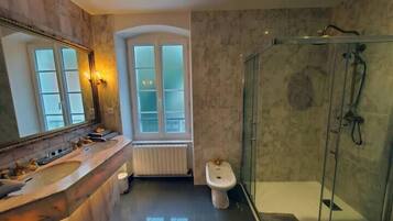 A bathroom with a marble vanity, a glass shower enclosure, a bidet, and a window with frosted glass panels.