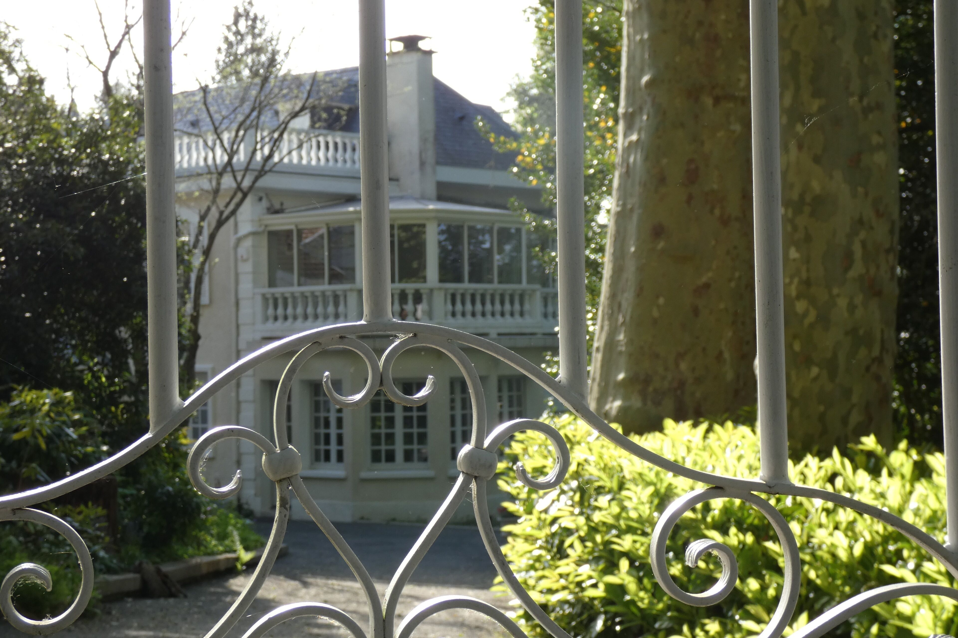 Une porte en fer forgé ornée d’une balustrade, un bâtiment blanc avec un balcon en arrière-plan, et un feuillage verdoyant.