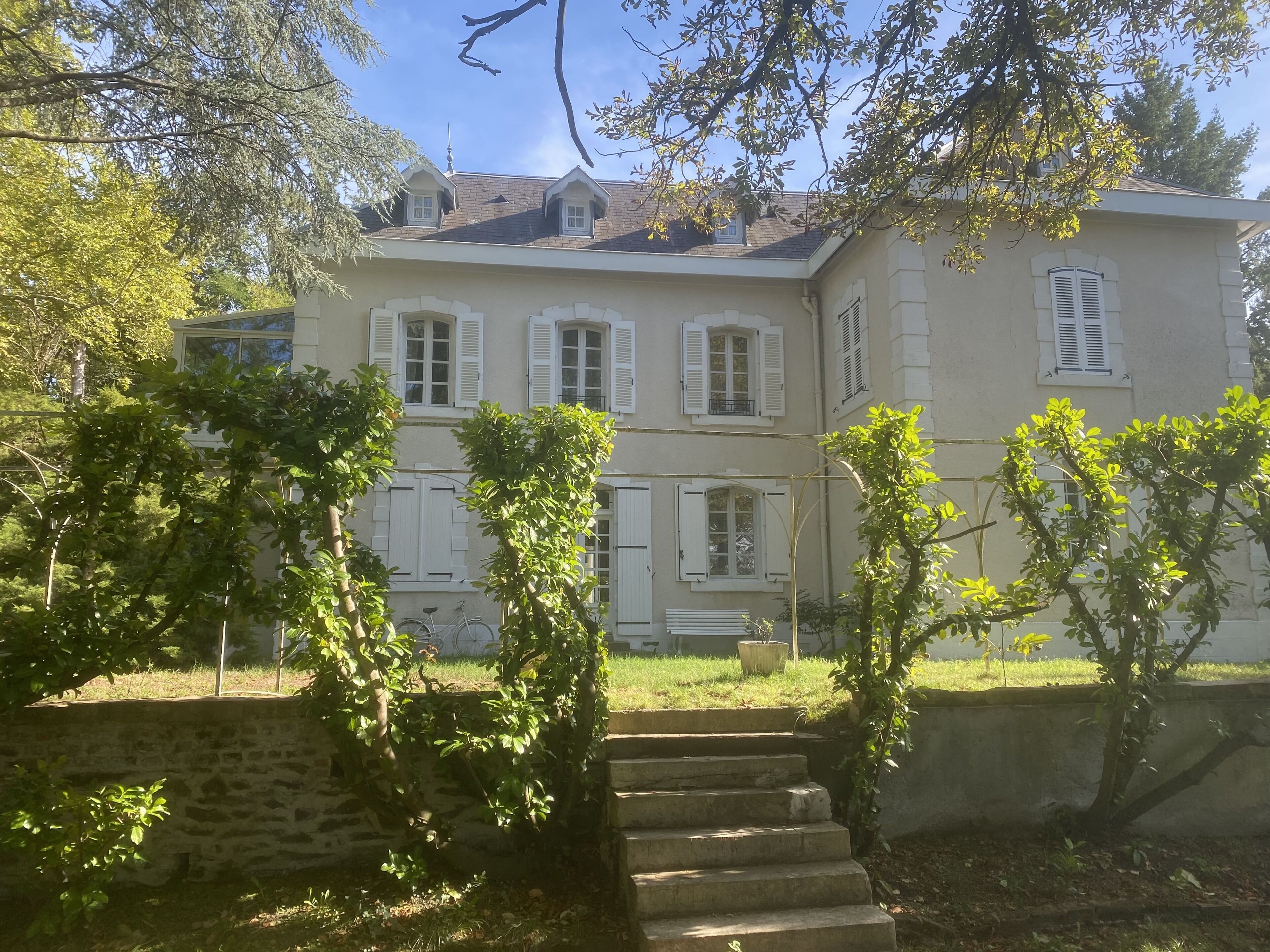 Ein zweistöckiges Haus mit weißen Fensterläden, einer Steintreppe und einem Garten mit Bäumen.
