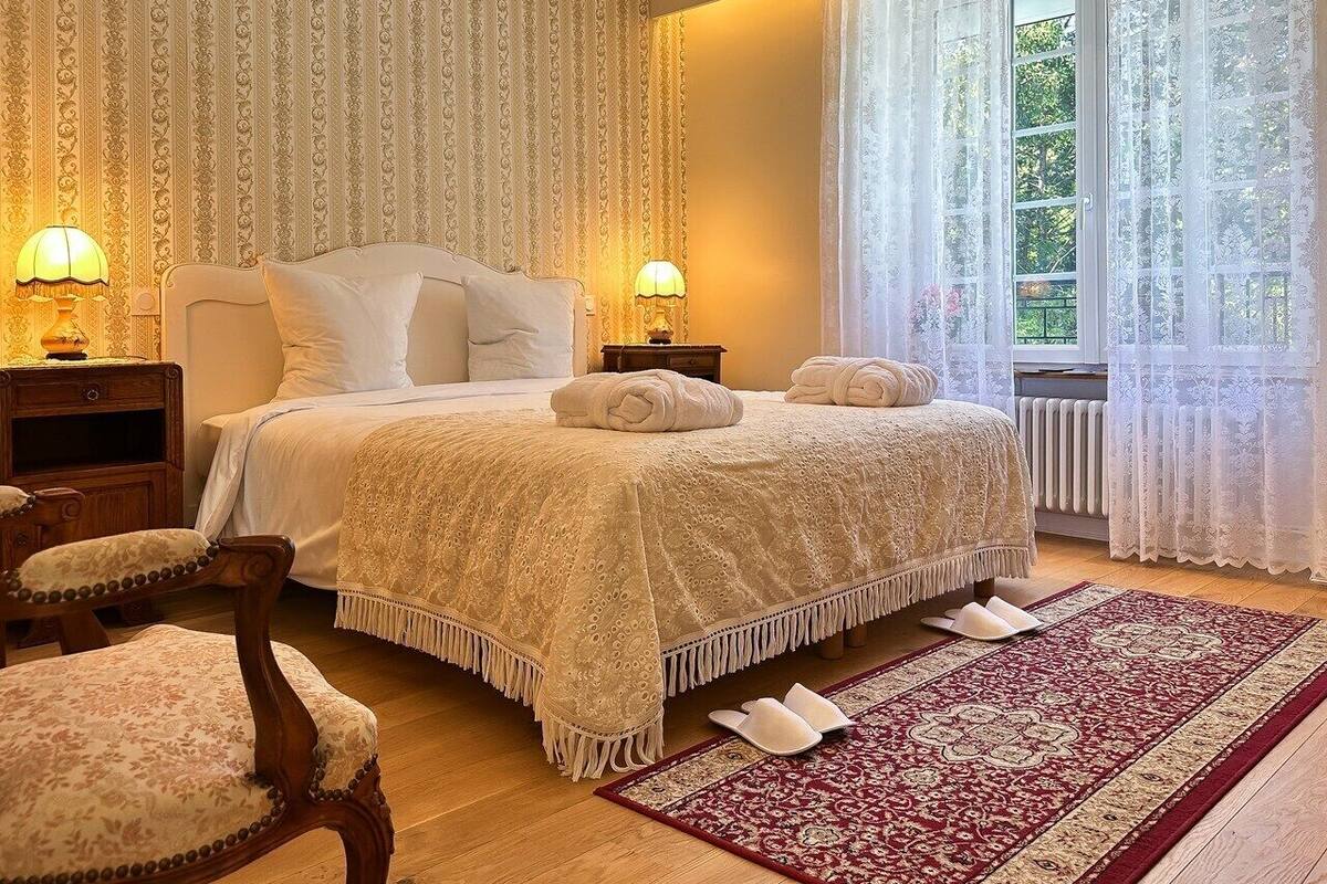 A bedroom with a large bed, two bedside tables with lamps, a wooden chair, a red and beige patterned rug, and a window with sheer curtains.