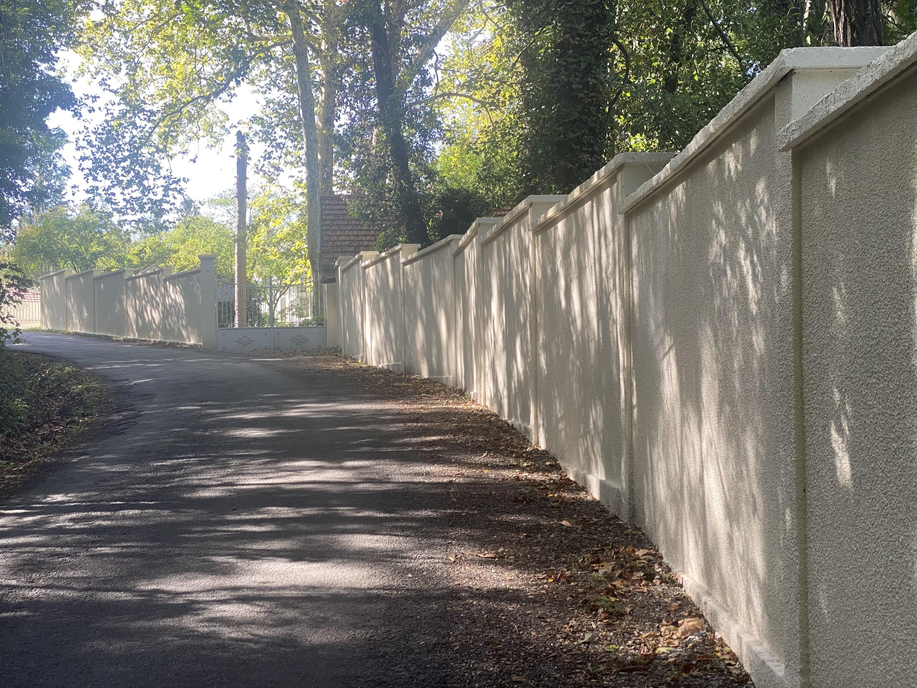 A paved pathway bordered by a high wall, with trees and sunlight filtering through.