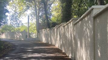 A paved pathway bordered by a high wall, with trees and sunlight filtering through.