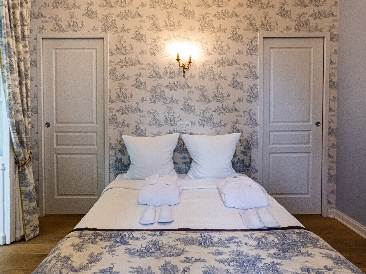 Una cama bien tendida con almohadas y toallas, un aplique de pared y una puerta con papel tapiz estampado con motivos florales.