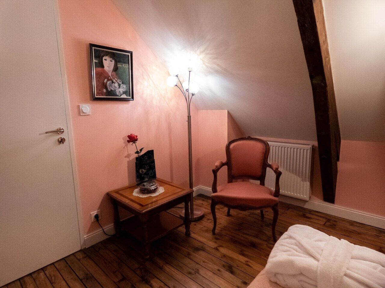 Ein Schlafzimmer mit Holzboden, einem rosafarbenen Sessel, einem Nachttisch mit Blumenvase, einem gerahmten Bild an der Wand und einer Heizung.