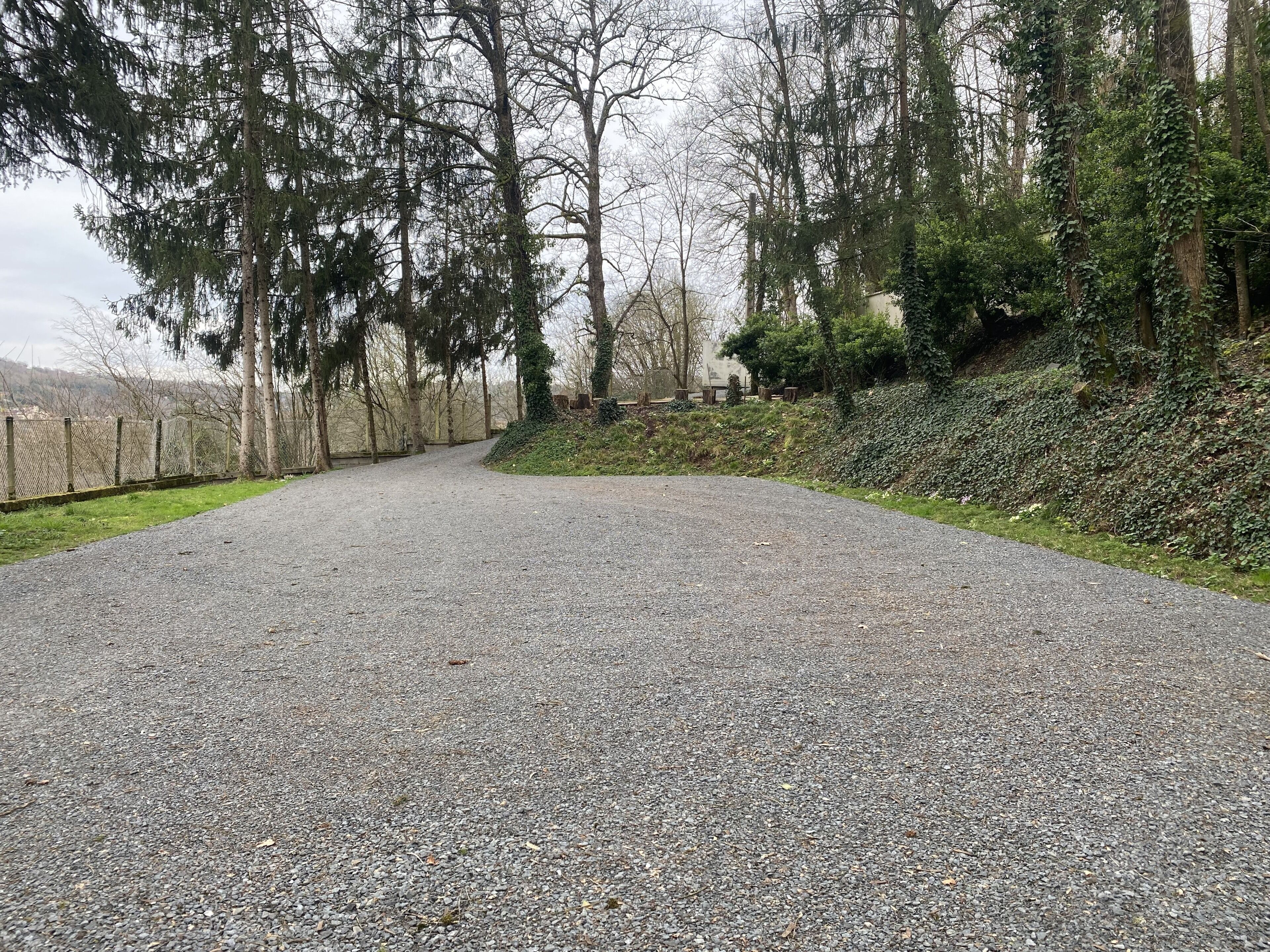 A gravel path in a wooded area with a chain-link fence on one side and a grassy slope on the other.