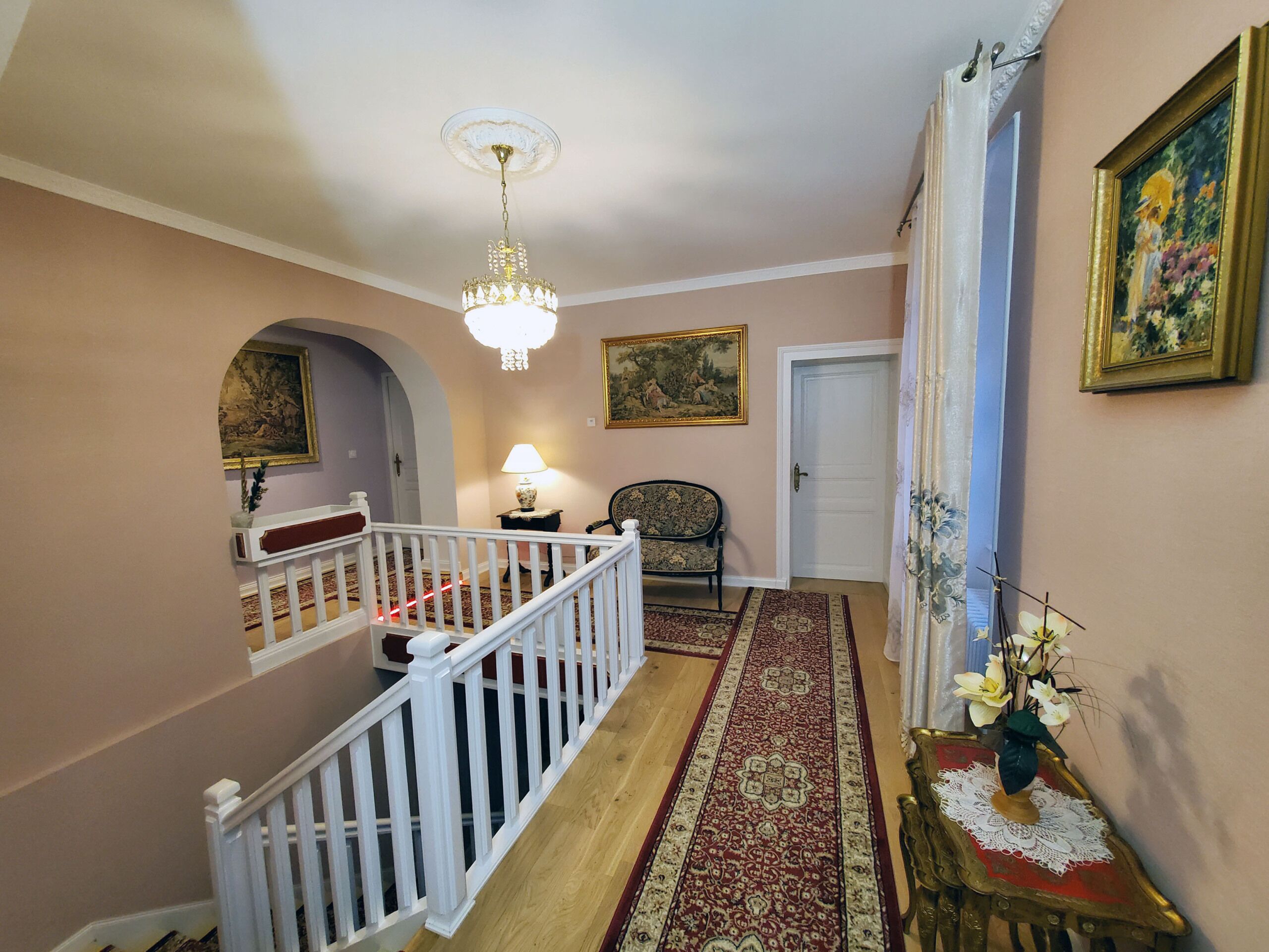 A hallway with a staircase, a chandelier, a painting, a lamp, and a vase with flowers.