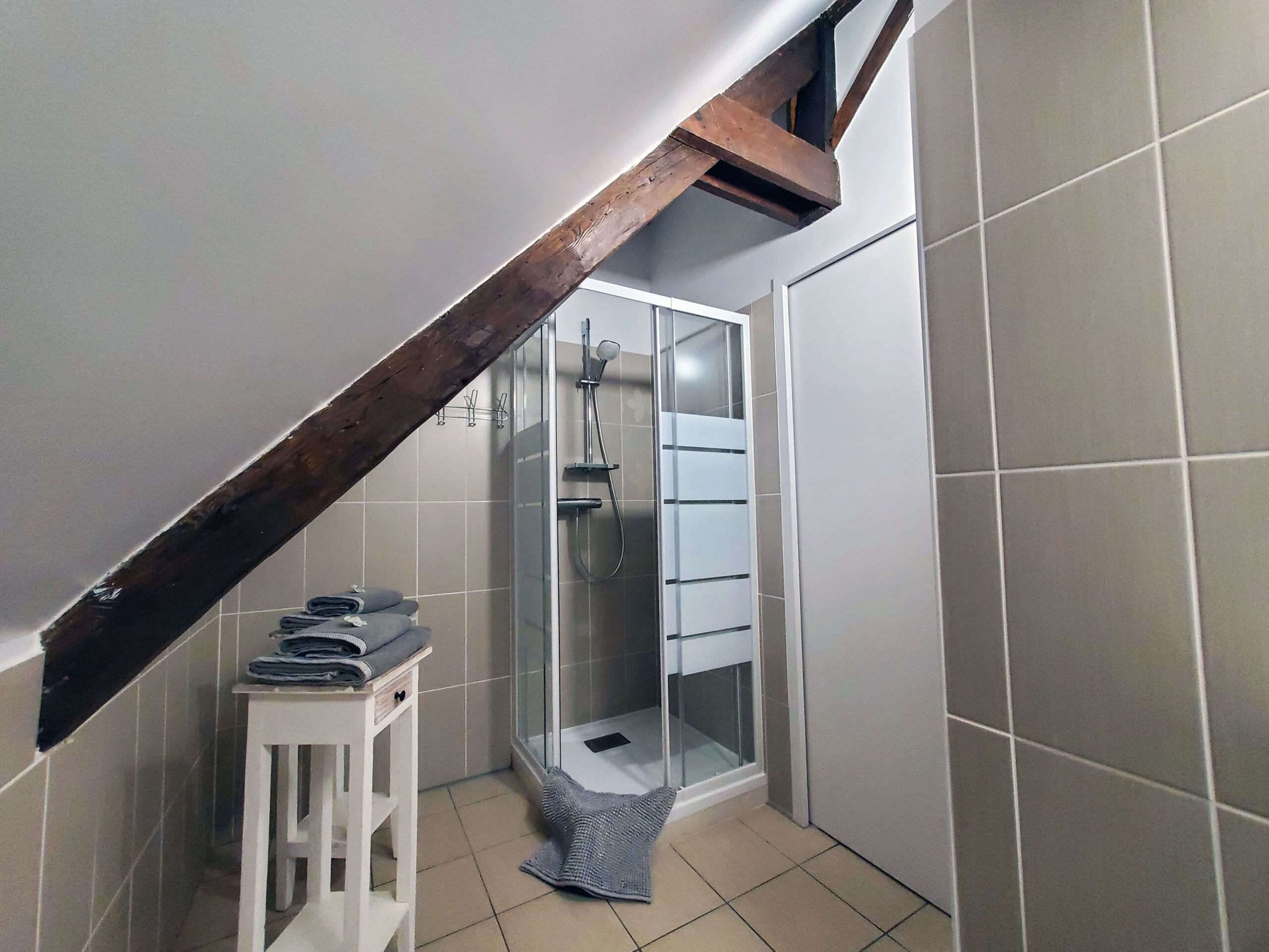 Un bagno con box doccia in vetro, uno sgabello bianco con asciugamani e travi in legno a vista.