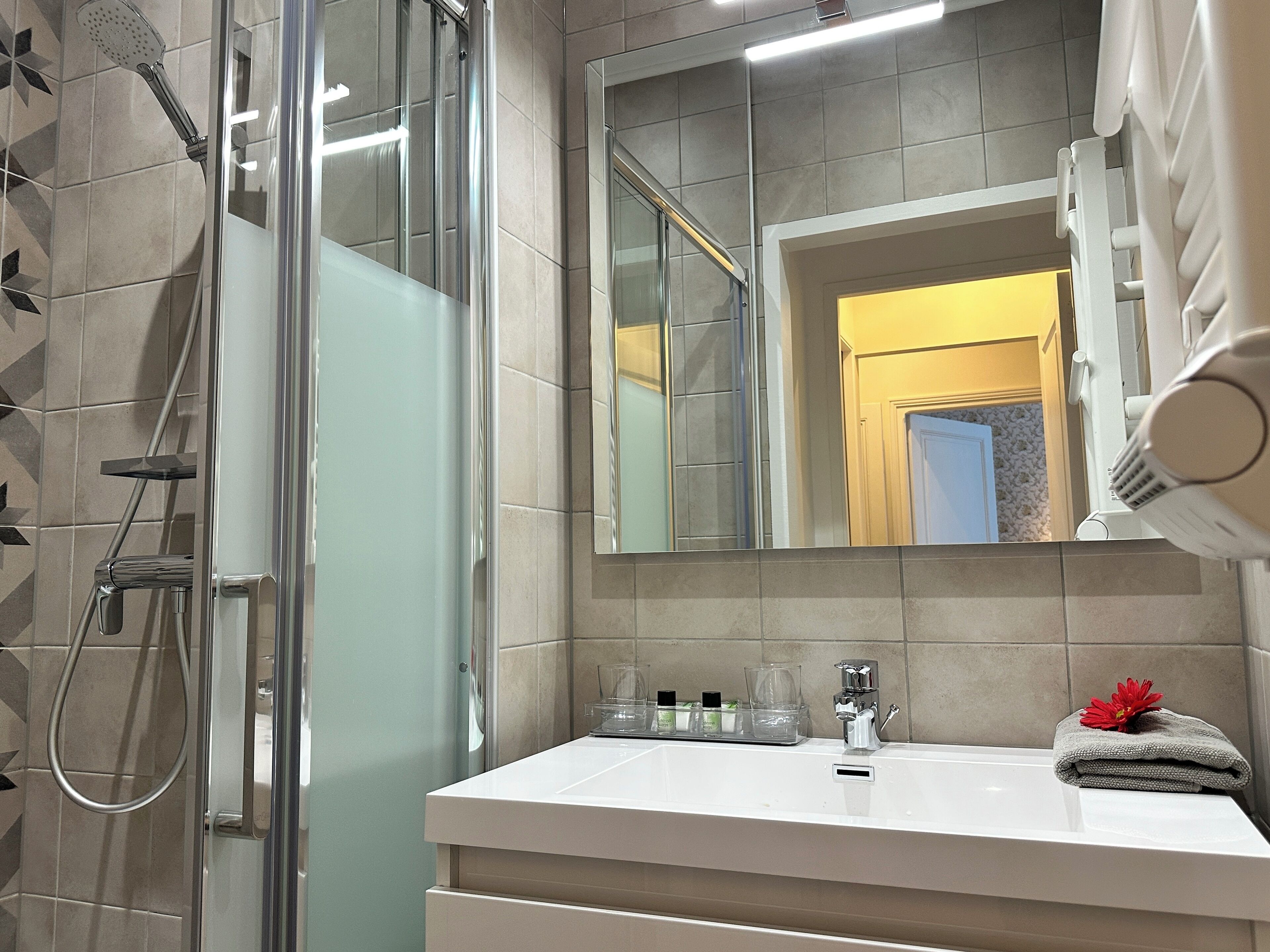 Un baño moderno con ducha envuelta en vidrio, un lavamanos blanco con un espejo encima y una toalla con un diseño floral en la encimera.