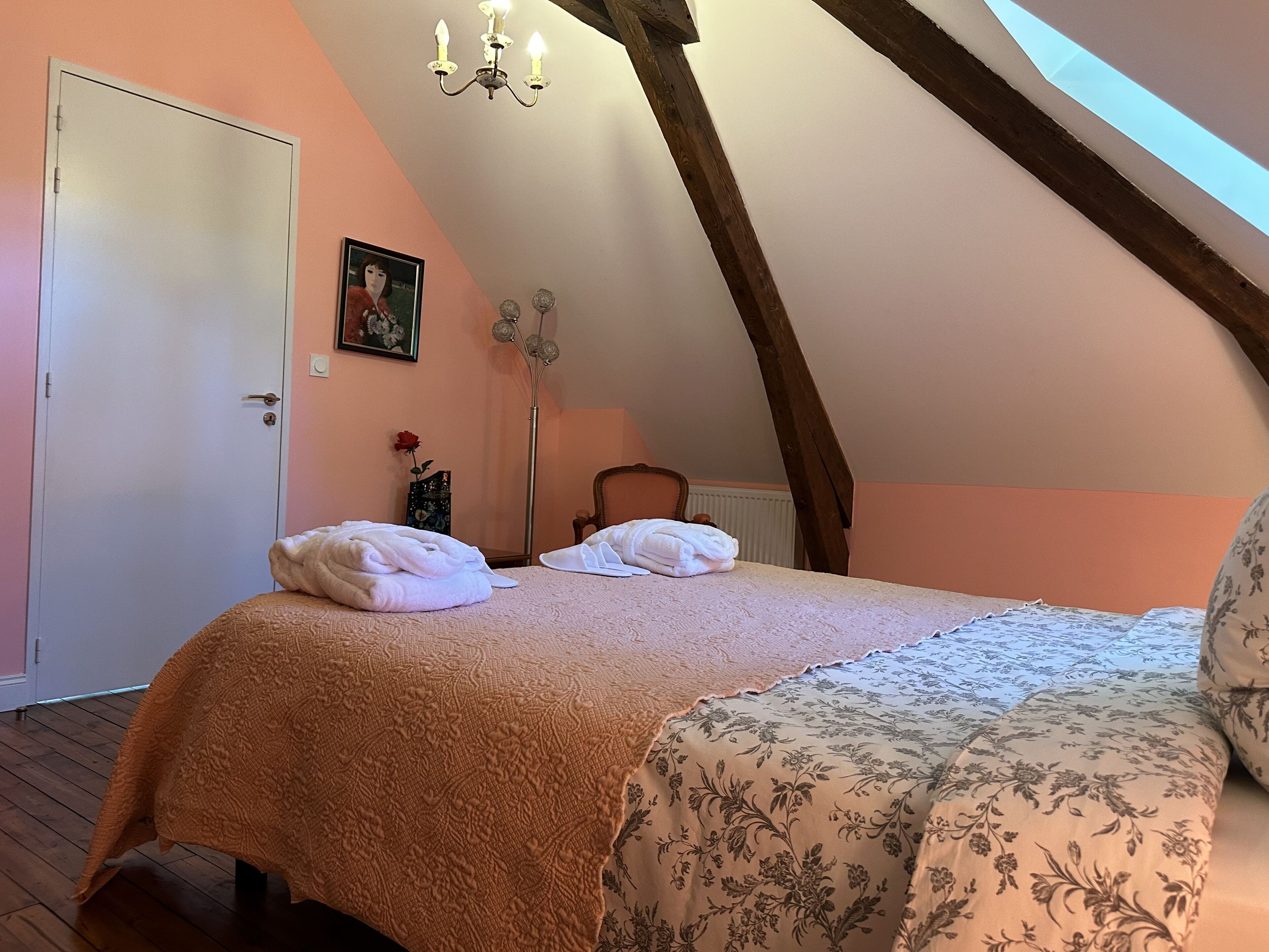 A bedroom with a sloped ceiling, a bed with a floral bedspread, a white door, a framed picture on the wall, and a tall floor lamp.