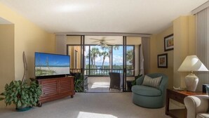Ein Wohnzimmer mit großem Fernseher, Deckenventilator und Blick auf Palmen durch große Fenster.