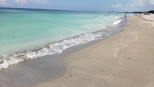 Ein Strand mit klarem türkisblauem Wasser, einem sandigen Ufer und ein paar Personen in der Ferne.